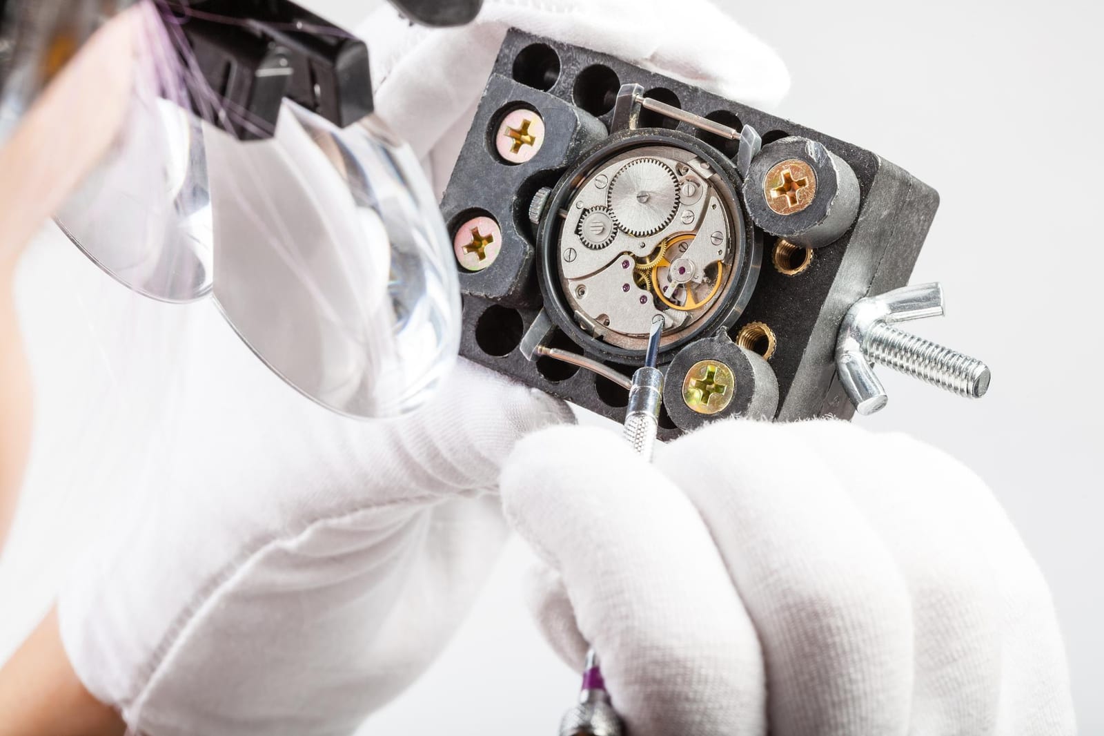 Mani con guanti bianchi e lente da orologiaio che regolano un piccolo movimento meccanico fissato in una morsa da banco